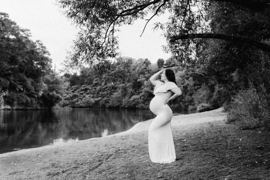 Black and white maternity photo of pregnant woman in white dress posing by a serene river surrounded by lush trees.