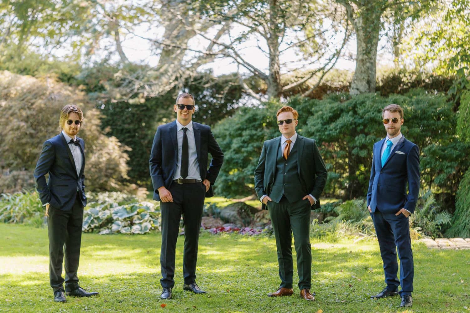 Four groomsmen in suits and sunglasses posing outdoors in a lush garden setting on a wedding day.