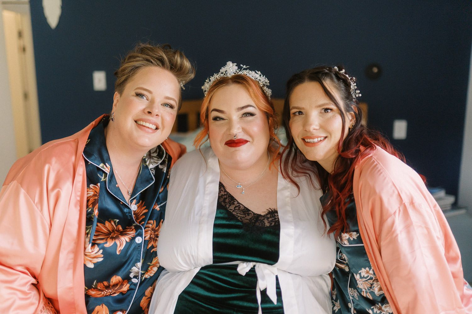 Three women in robes and floral pajamas smile together during bridal getting-ready preparations.