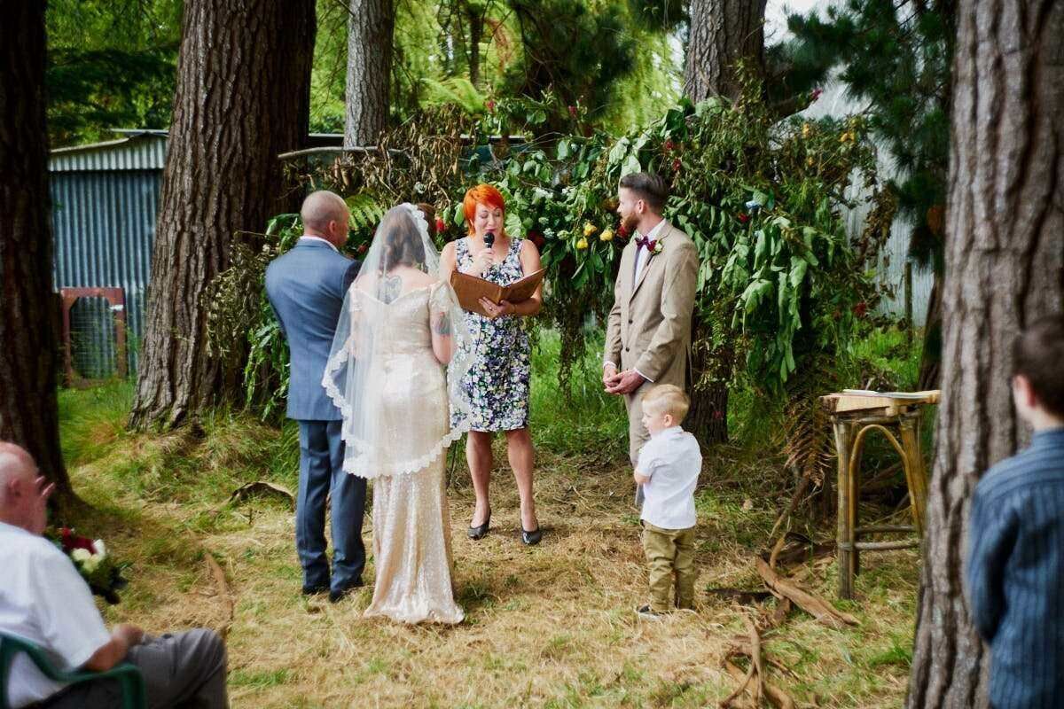 Outdoor wedding ceremony in a forest setting with bride, groom, and officiant standing among tall trees.