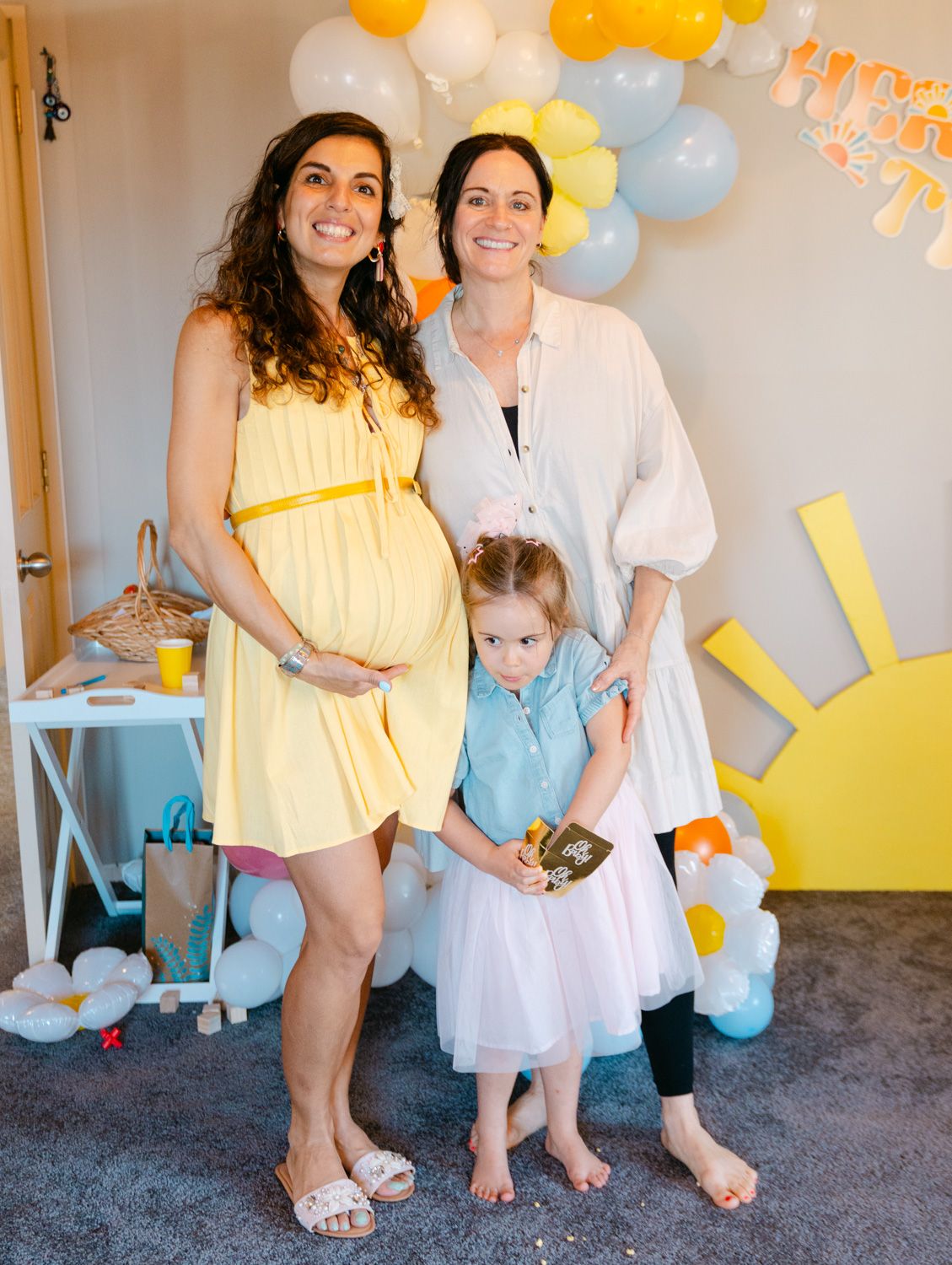 Two women and a young girl pose at a sunny baby shower with balloon decorations.