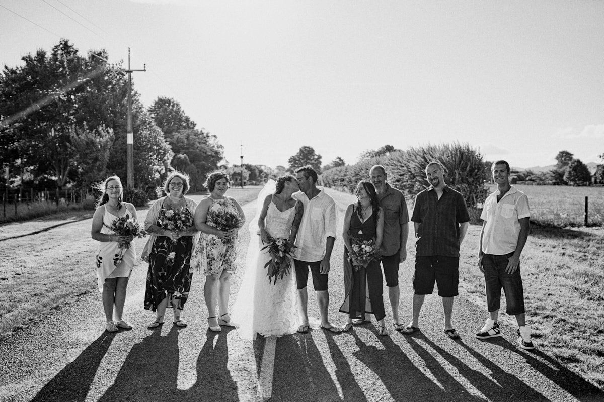 Black and white wedding photo of bride, groom, and guests posing together on a sunny rural road.