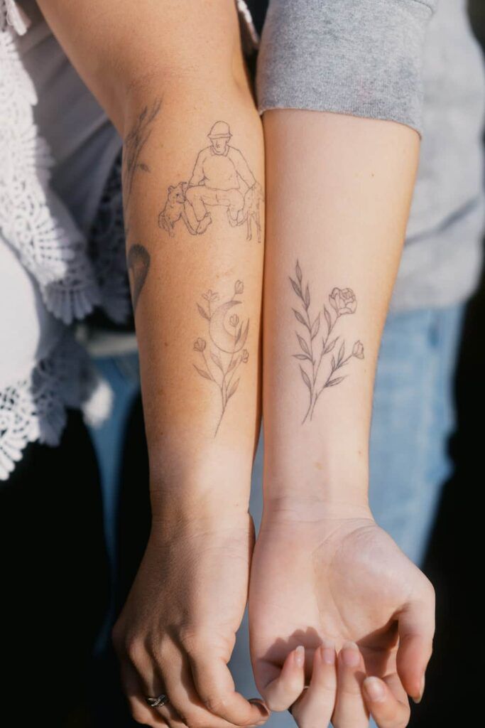 Two people display matching fine line floral tattoos on their forearms, with delicate botanical designs inked in black.