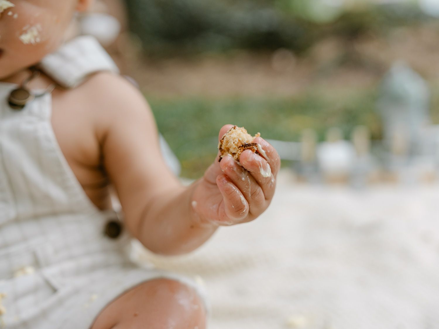 A baby in linen overalls sits outdoors, holding a small piece of food in their chubby hand.