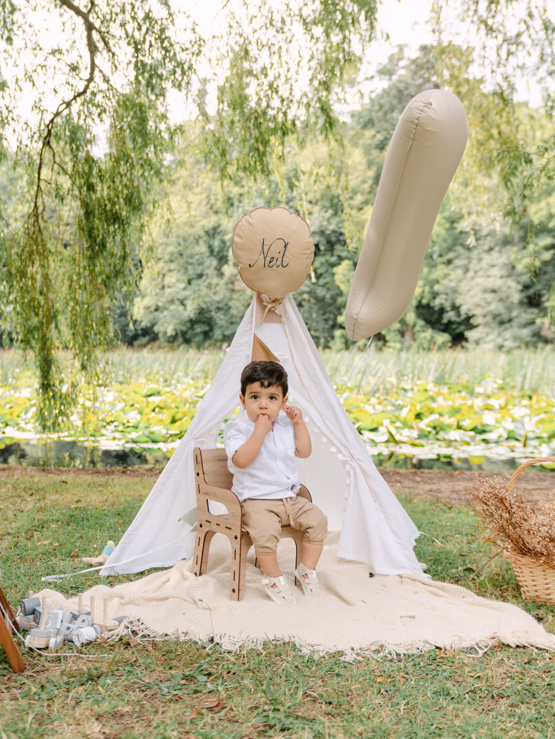 Toddler boy named Neil sits in a wooden chair inside a white teepee outdoors, surrounded by neutral balloons and greenery.