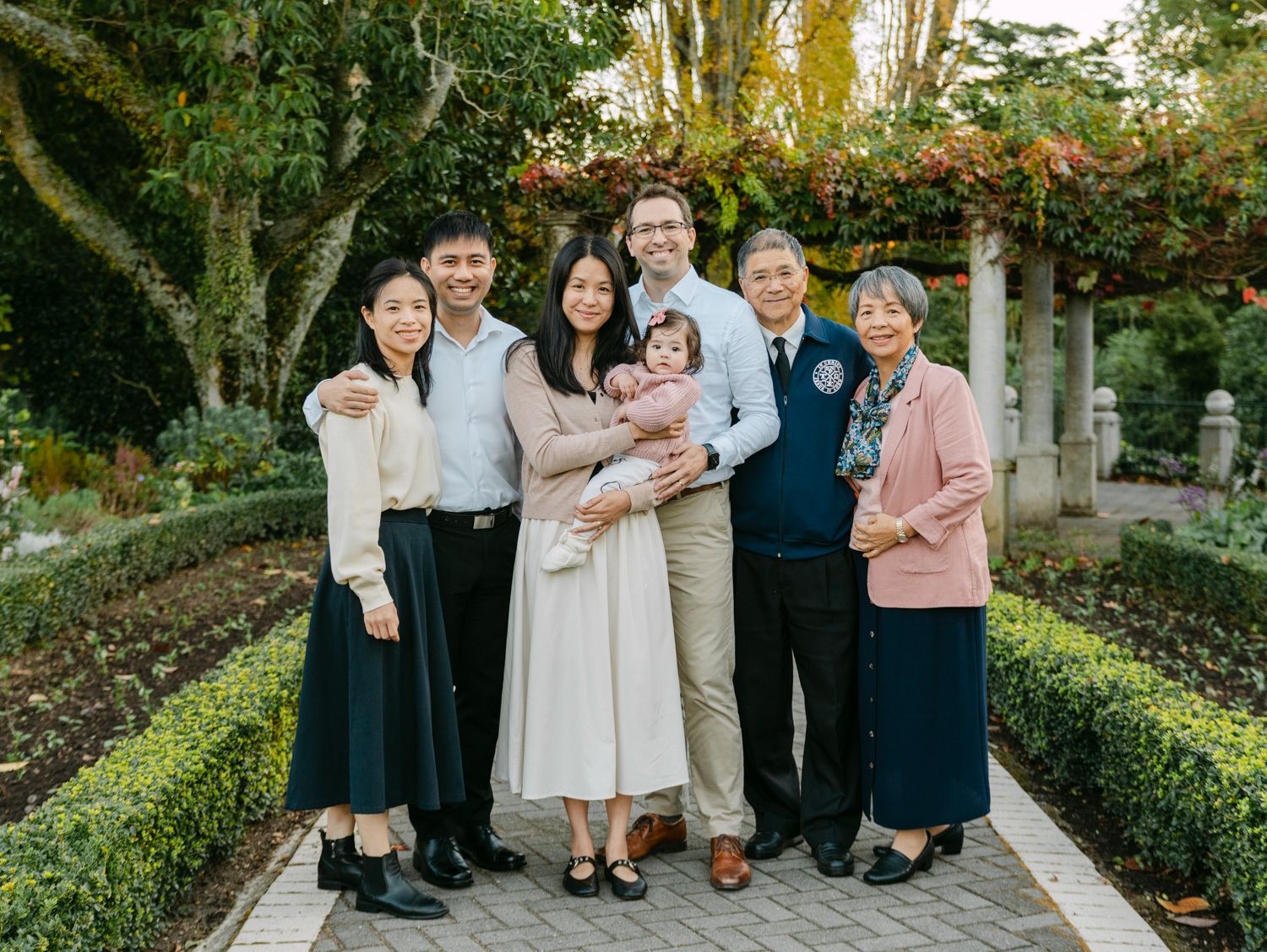 Multi-generational family of seven smiling together in a lush garden pathway, including a baby in white.