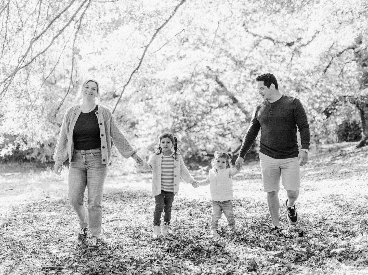 Black and white photo of a family of four holding hands and walking through a leaf-covered park on a sunny day.