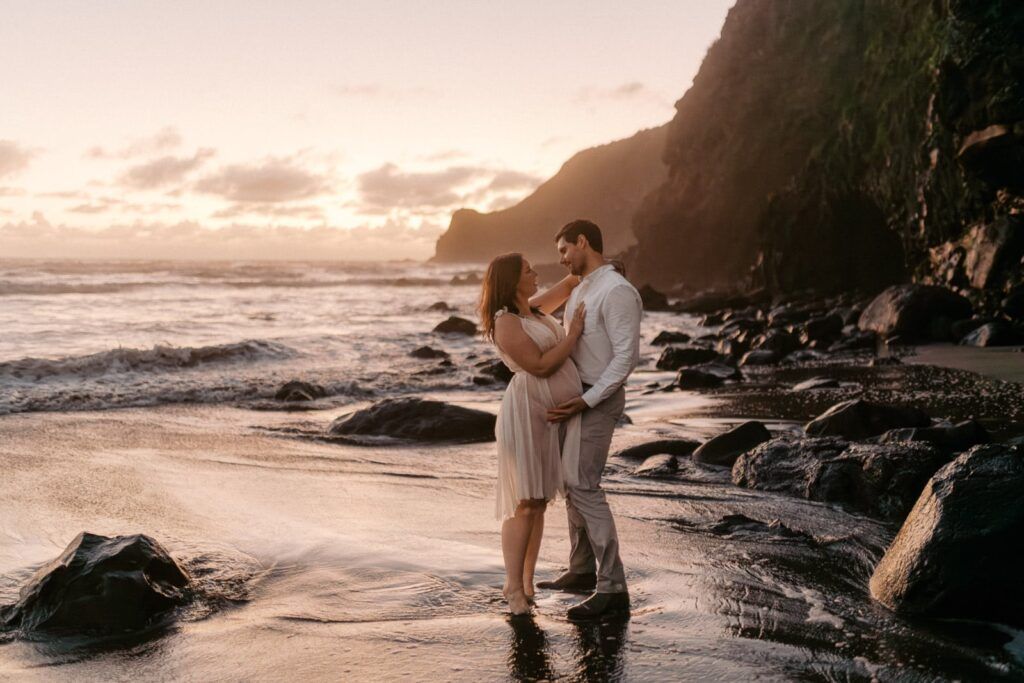 Couple embracing on a rocky beach at golden sunset with ocean waves and dramatic cliffs in the background.