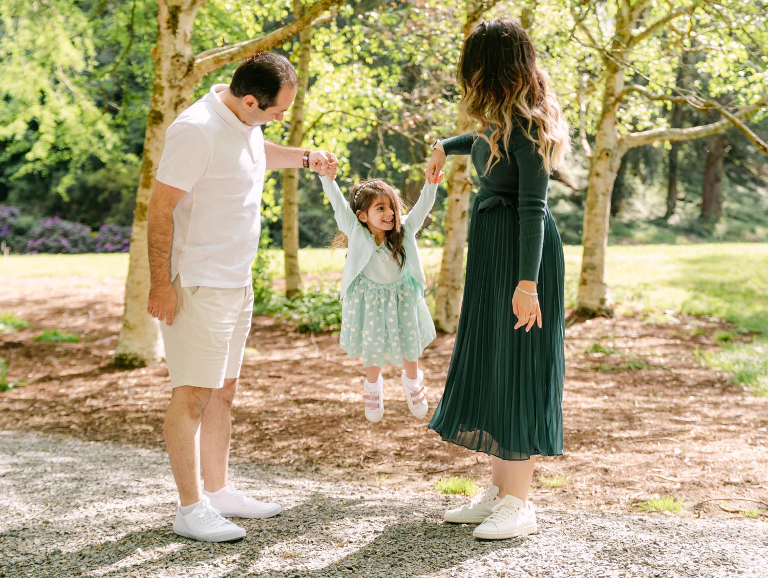 Parents swing their laughing little girl between them outdoors in a sunlit park setting.