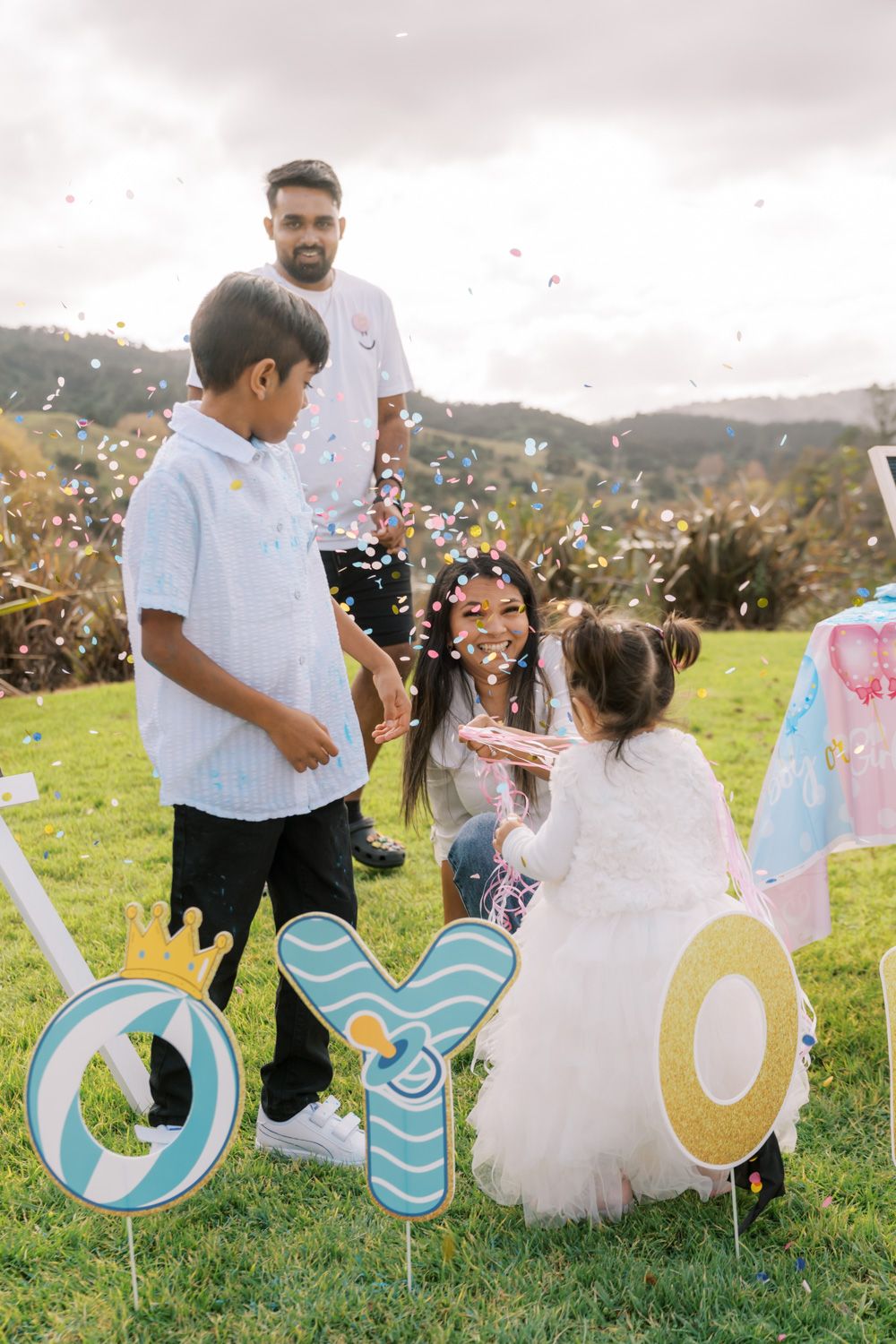 Family celebrating a birthday outdoors with colorful confetti, decorative OYO letters on grass, surrounded by green hills.