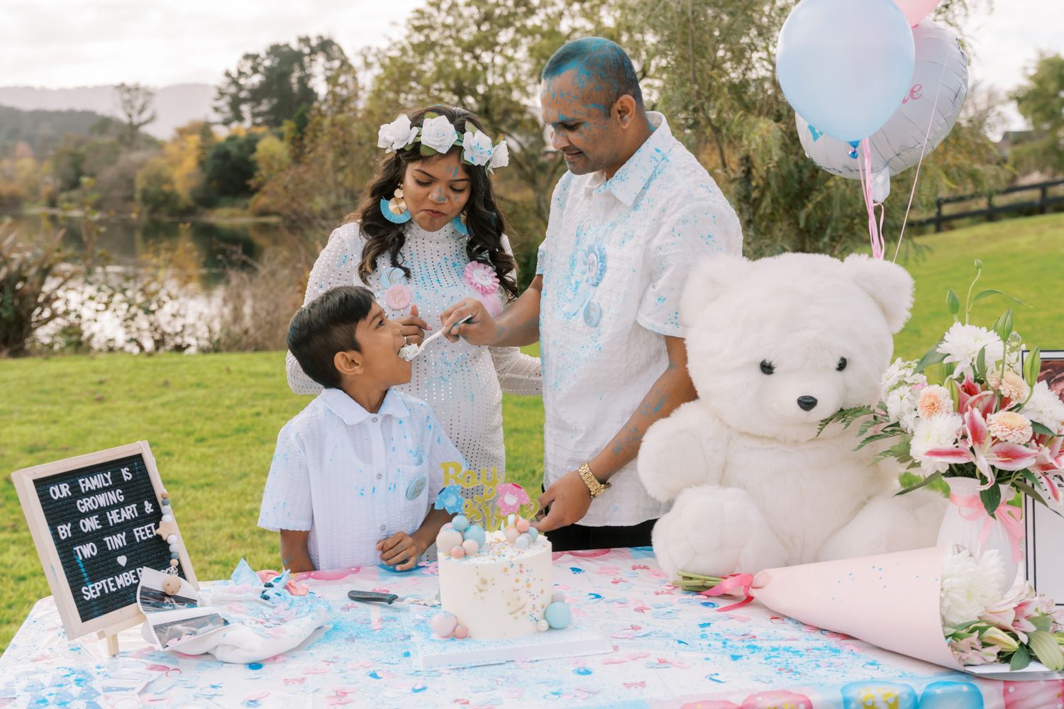 Family celebrating a gender reveal party outdoors by a lake with a large white teddy bear and pink and blue decorations.