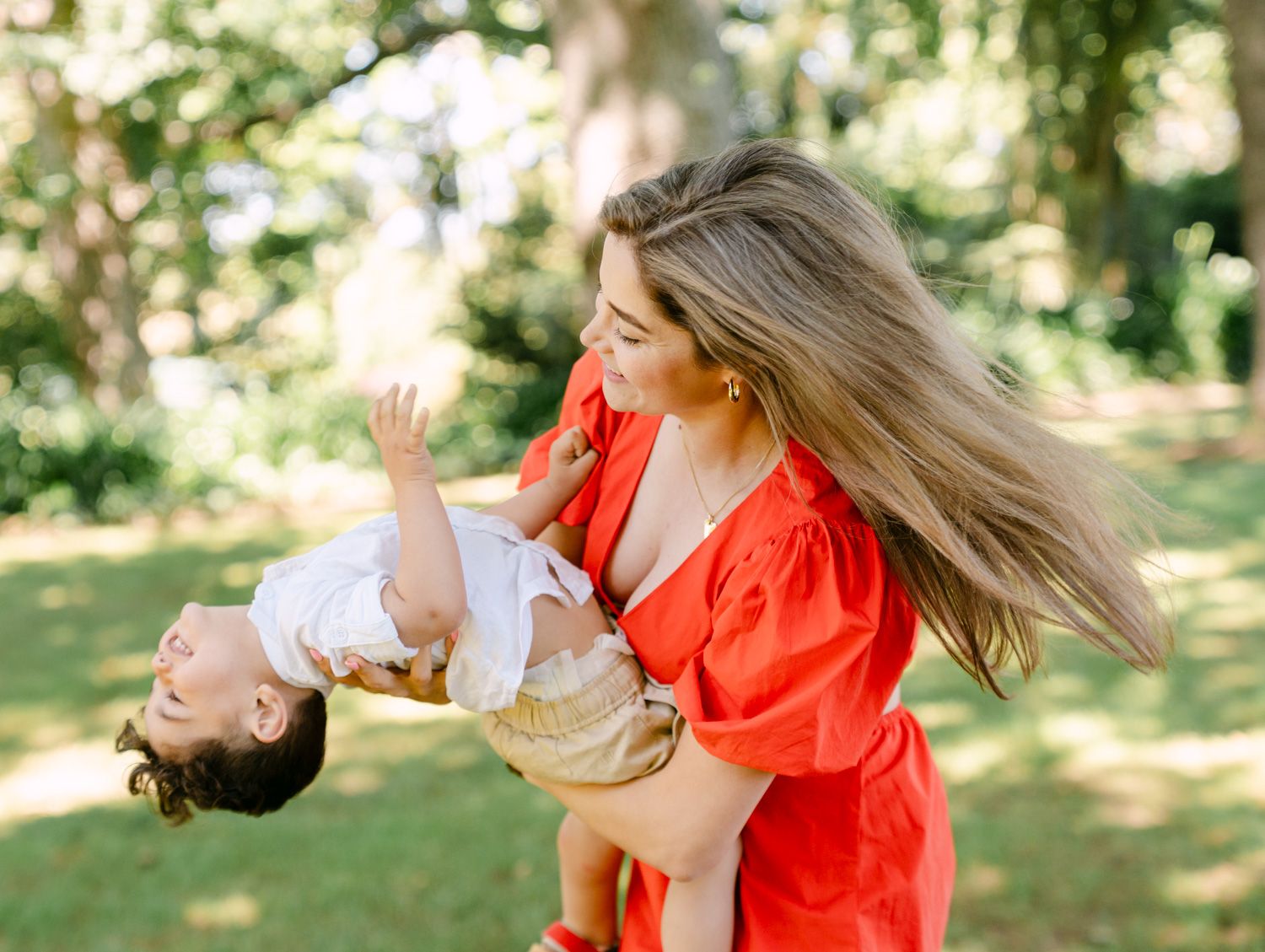A joyful mother in a red dress playfully swings her laughing toddler upside down in a lush green park.