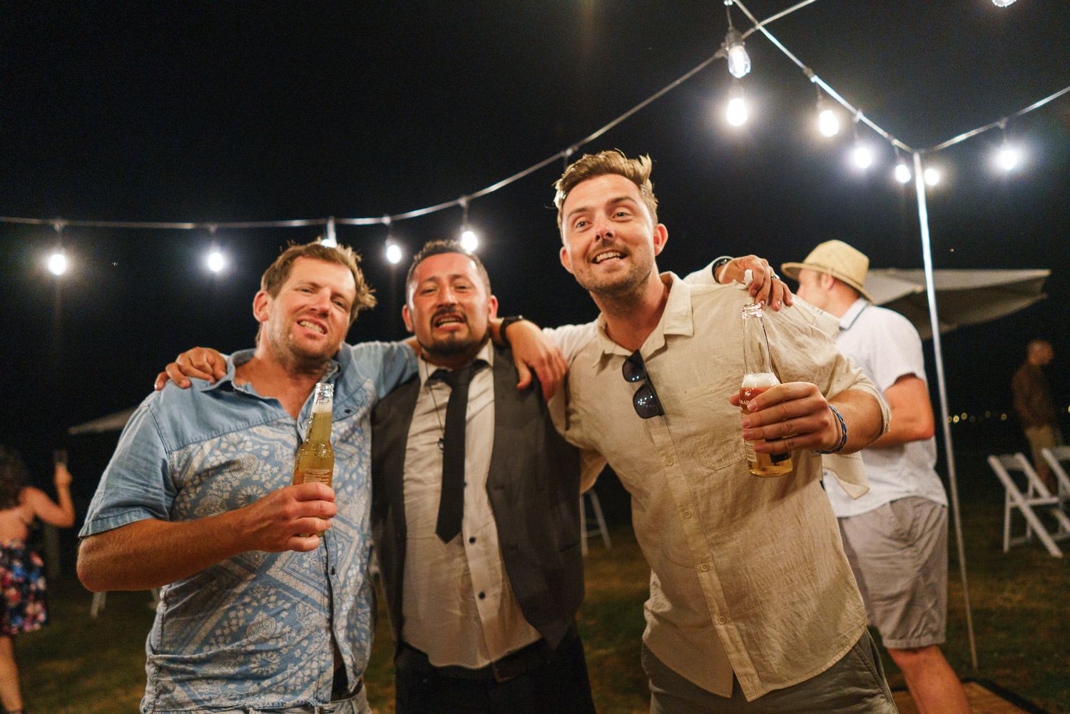Three men smiling and posing together at an outdoor night event with string lights.