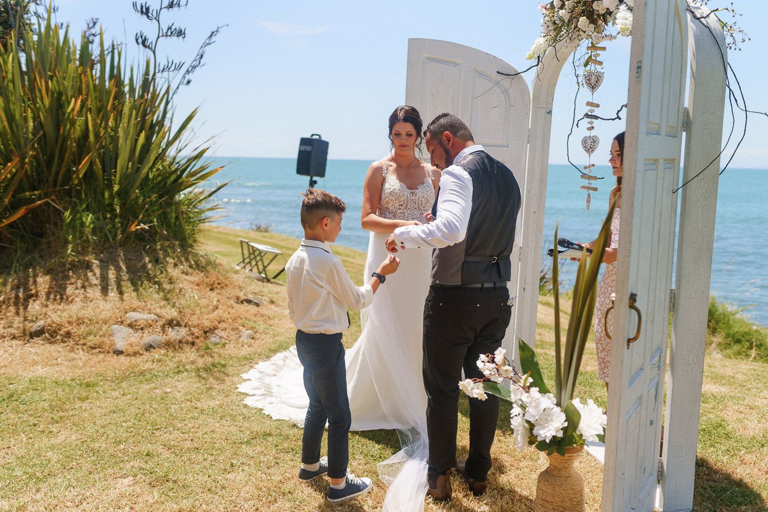 Outdoor beach wedding ceremony with bride, groom, and young boy standing beneath a white door arch by the ocean.