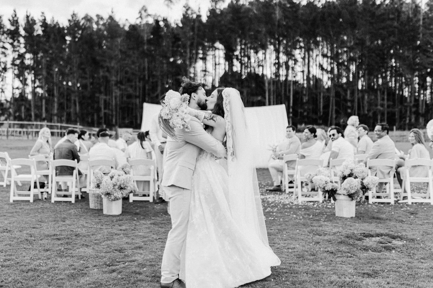 Newlyweds share romantic kiss on lawn at outdoor wedding reception surrounded by guests and pine trees.
