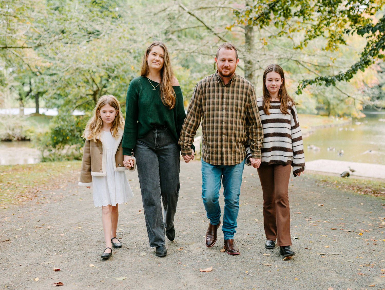 Family of four walking hand-in-hand on a park path surrounded by autumn trees near a pond.