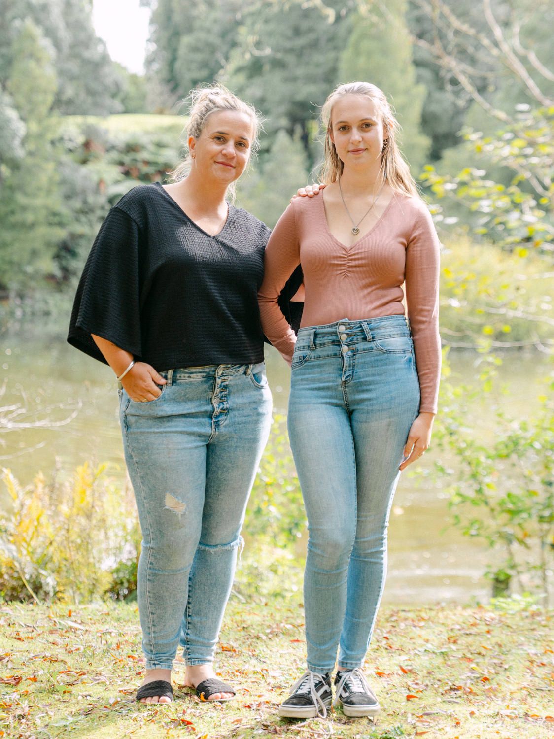 Two young women in jeans pose together outdoors near a pond, one in black top, one in mauve long-sleeve shirt.