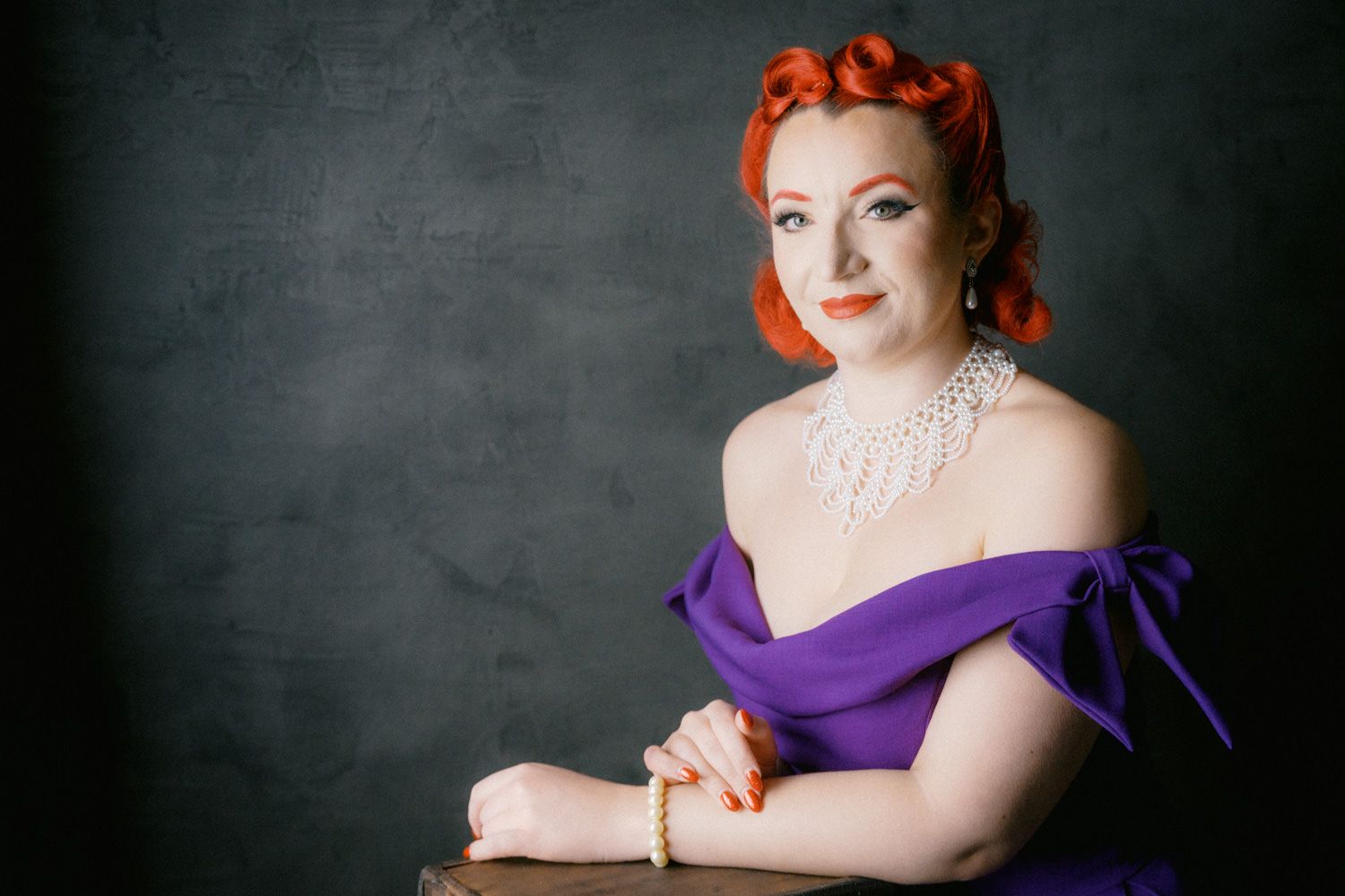 Woman with red hair in purple off-shoulder gown wearing pearl necklace poses elegantly against dark gray backdrop.