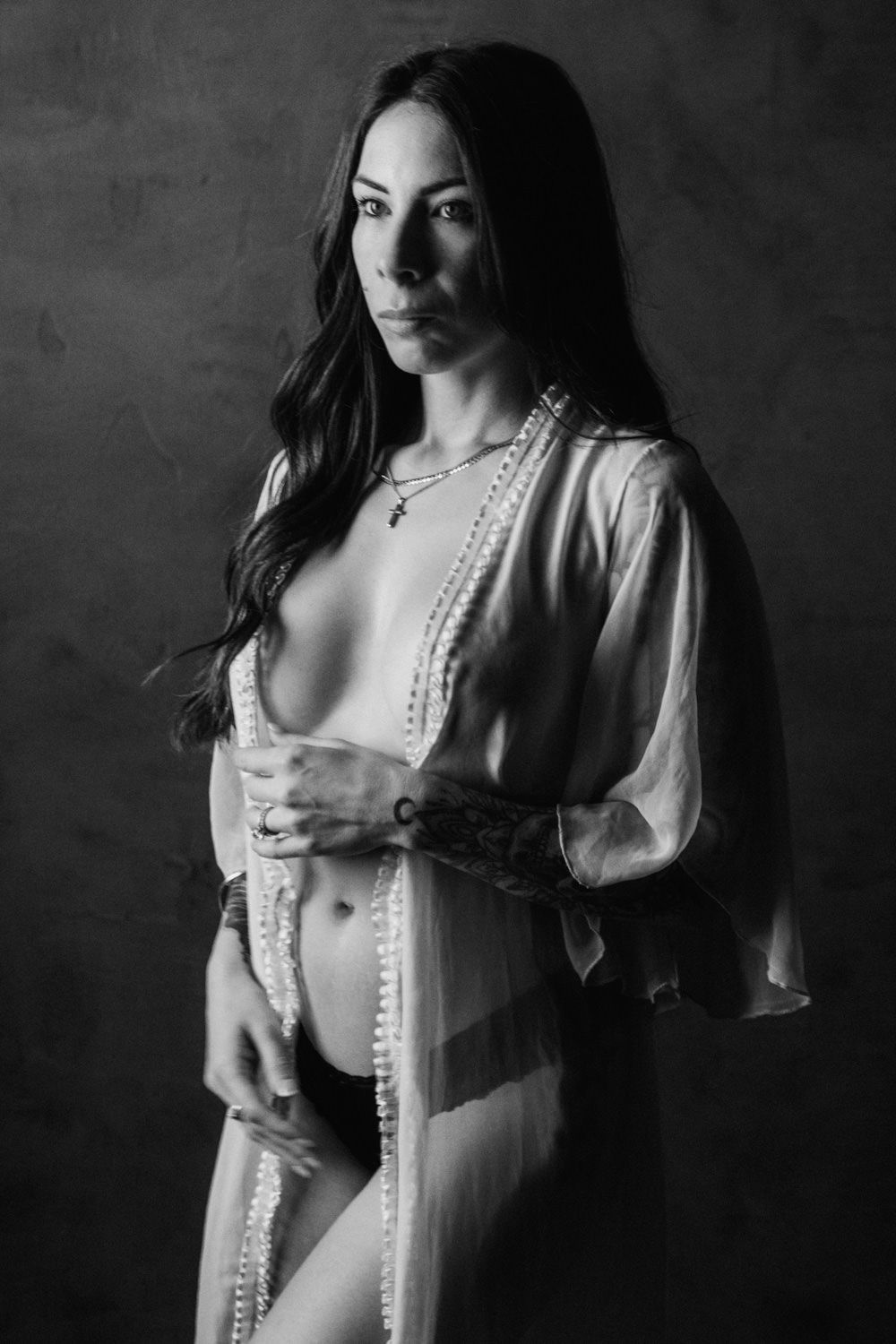Black and white portrait of woman in sheer robe with tattoo posing in studio.