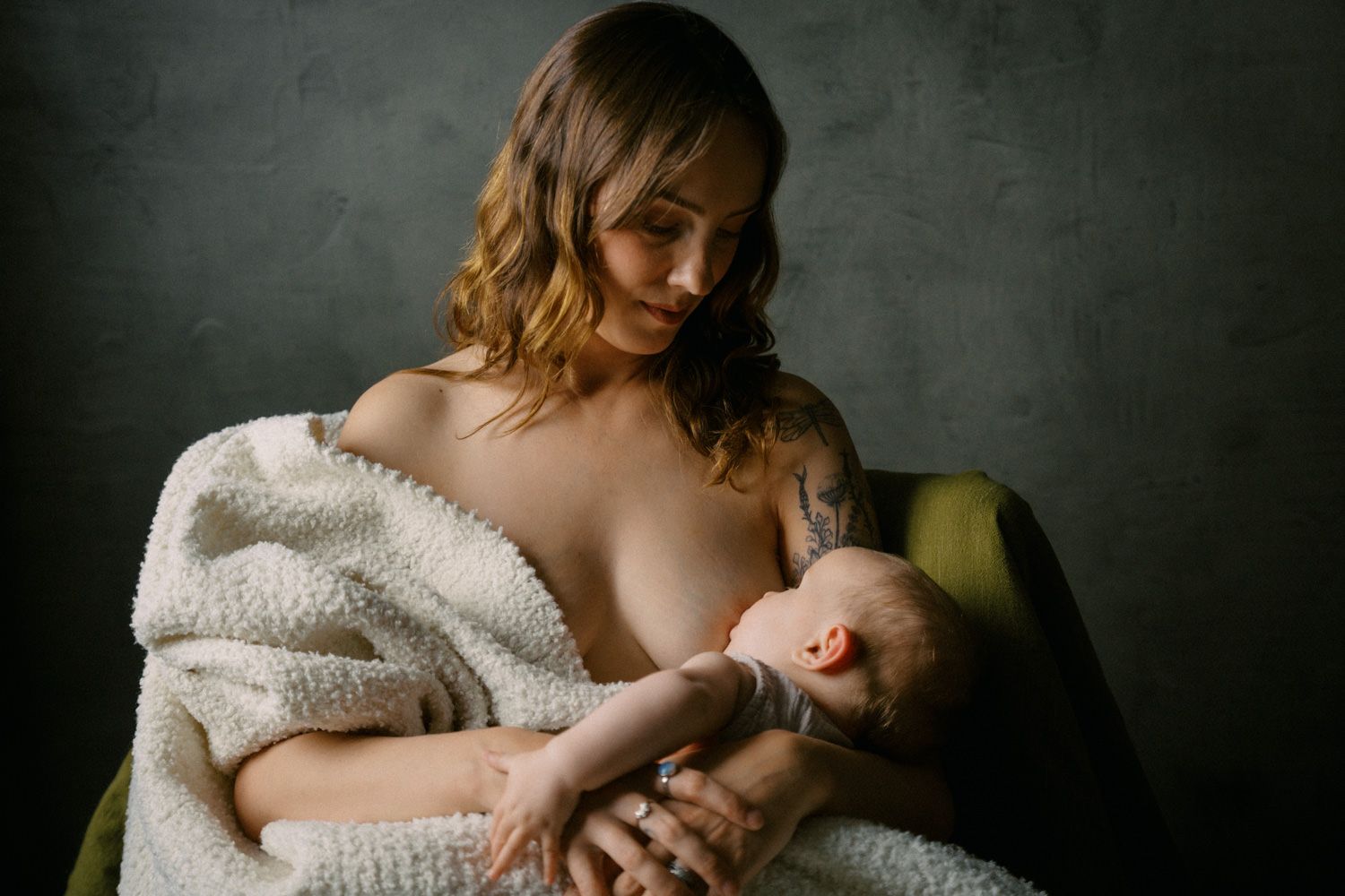 Mother tenderly breastfeeding her newborn baby while seated in a chair, wrapped in a soft white blanket.