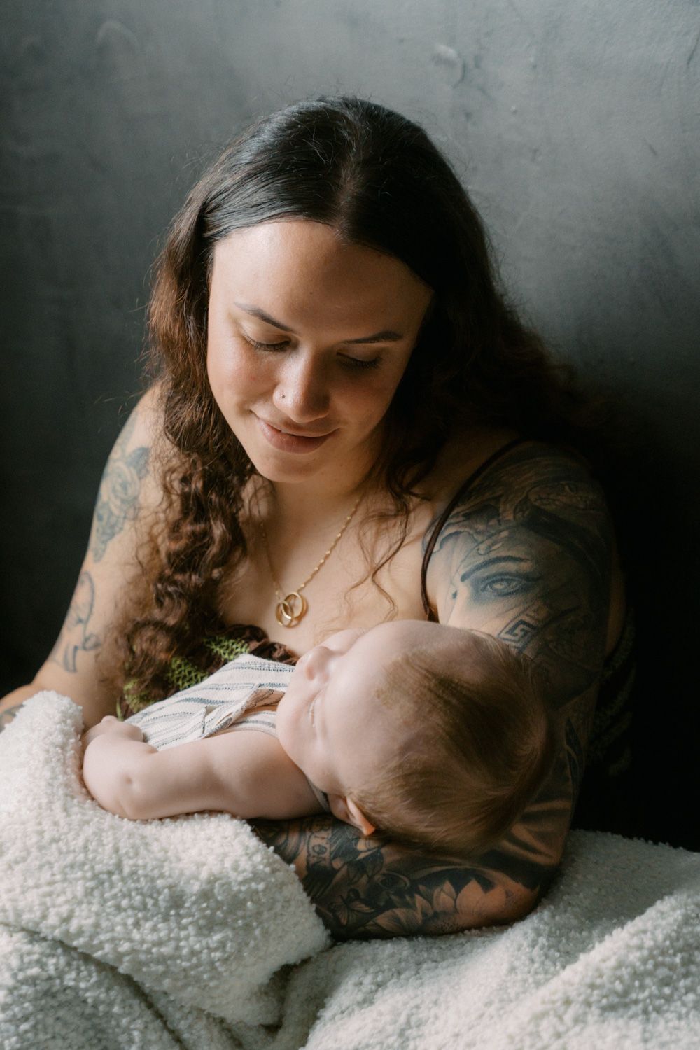 Tattooed mother tenderly breastfeeding her newborn baby wrapped in a white blanket against a dark wall.