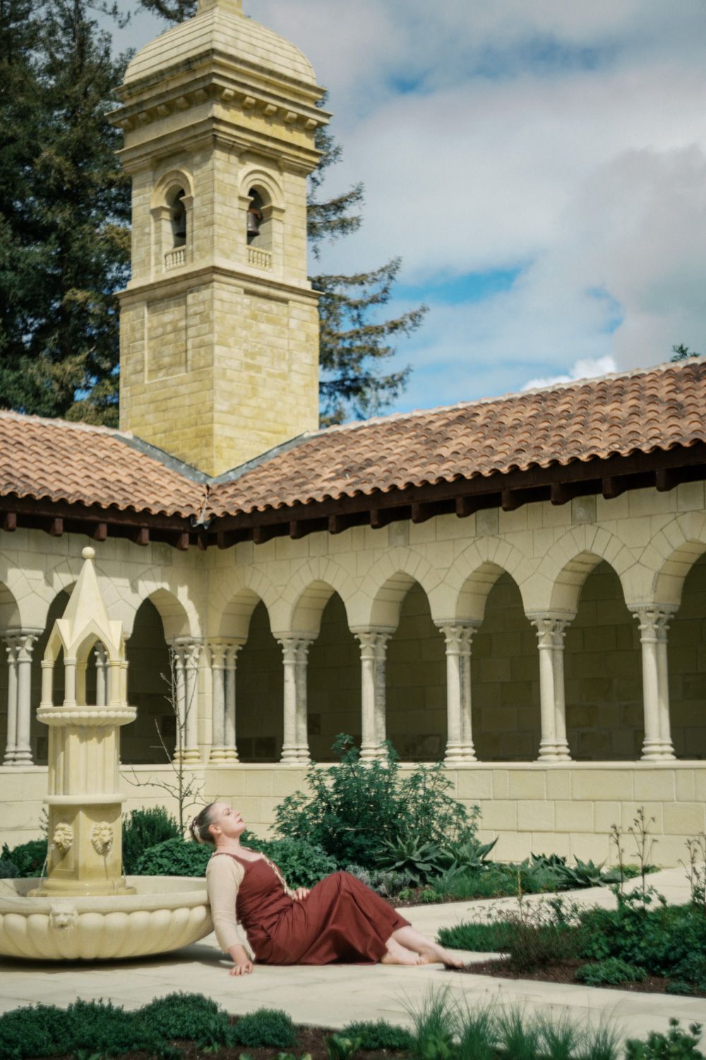 Woman in red dress reclines gracefully in a sunlit courtyard with arched colonnade and stone bell tower.