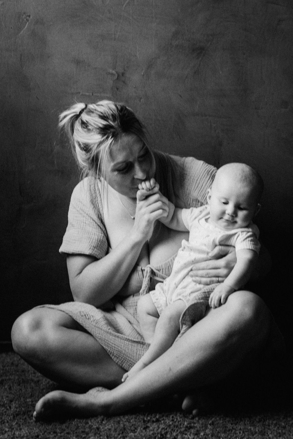 Black and white photo of a blonde mother sitting cross-legged on floor, tenderly holding her smiling newborn baby.