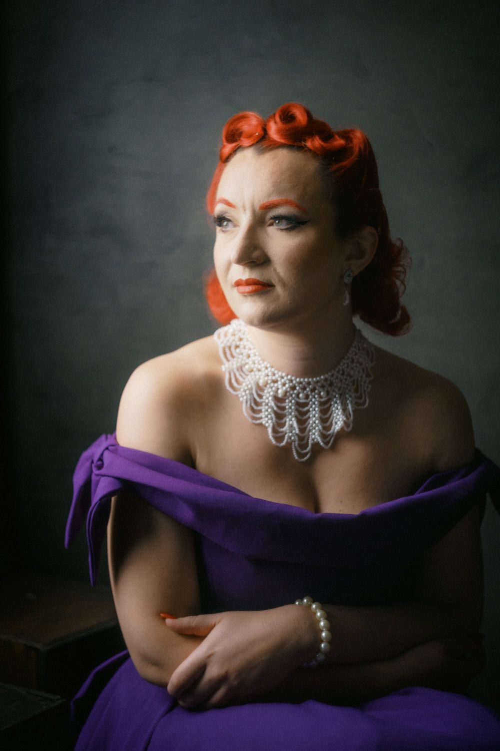 Woman with red pin-up hair wearing purple off-shoulder gown and pearl necklace poses elegantly against dark background.