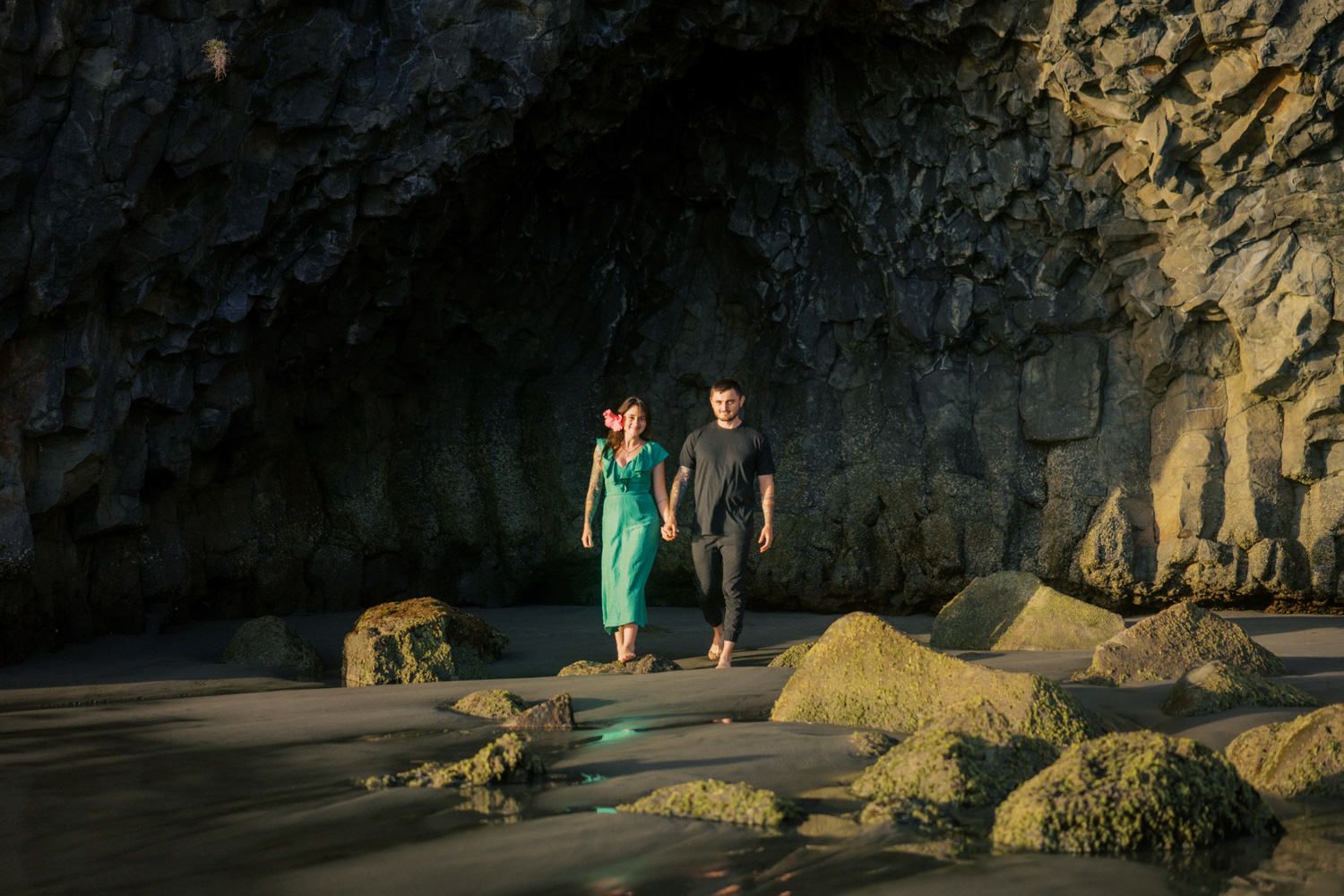 Couple walking hand in hand on a sandy beach near dramatic rock cave formations at golden hour.