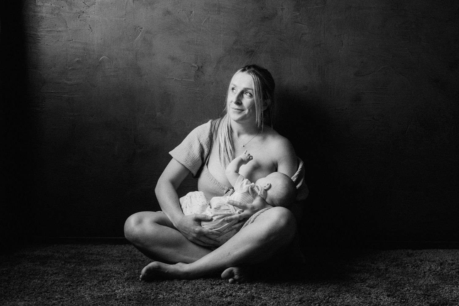 Black and white photo of a mother sitting cross-legged on floor, breastfeeding her newborn baby against dark background.