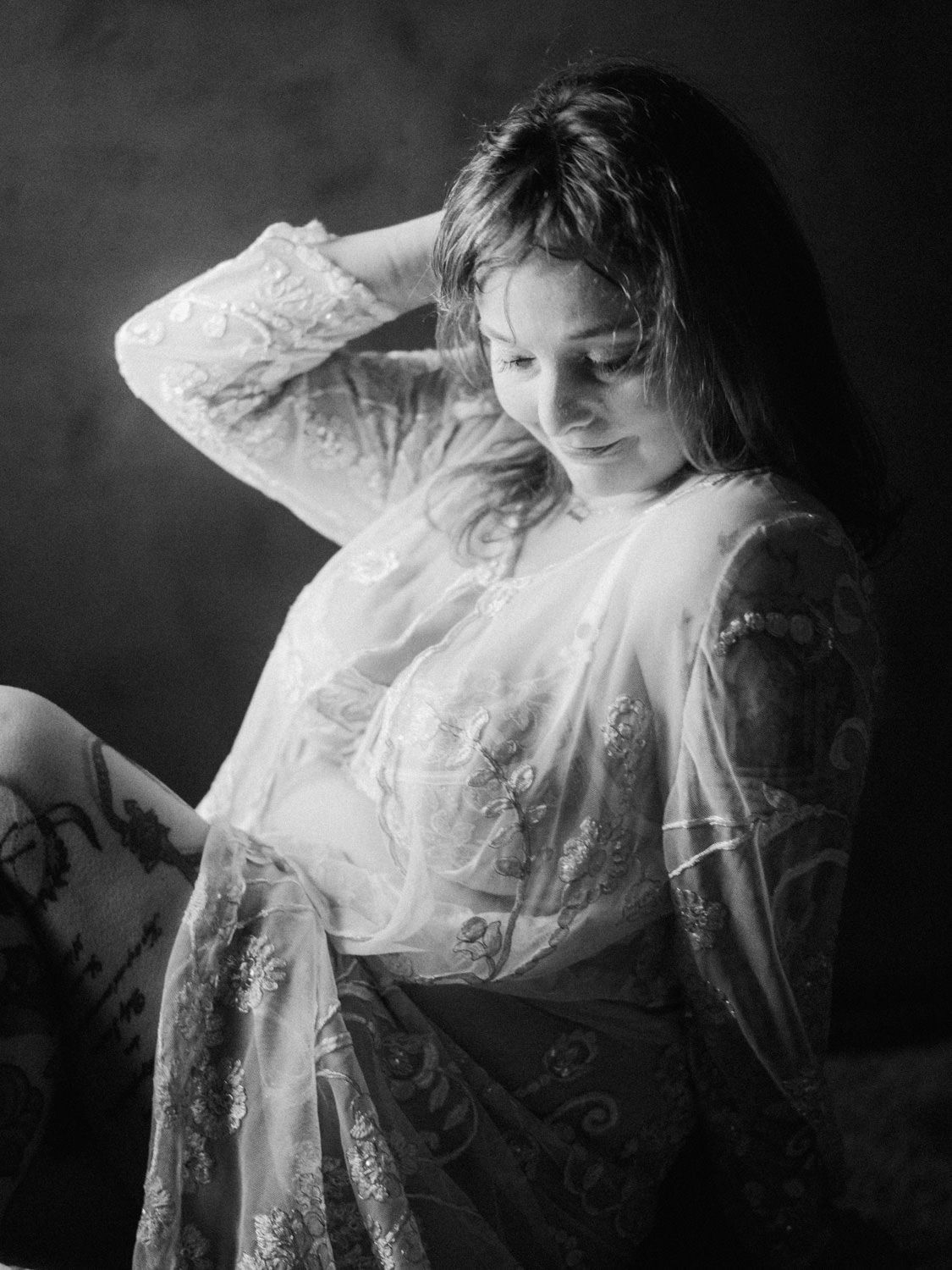 Black and white portrait of a woman in sheer lace robe, posing gracefully with hand in hair in studio lighting.