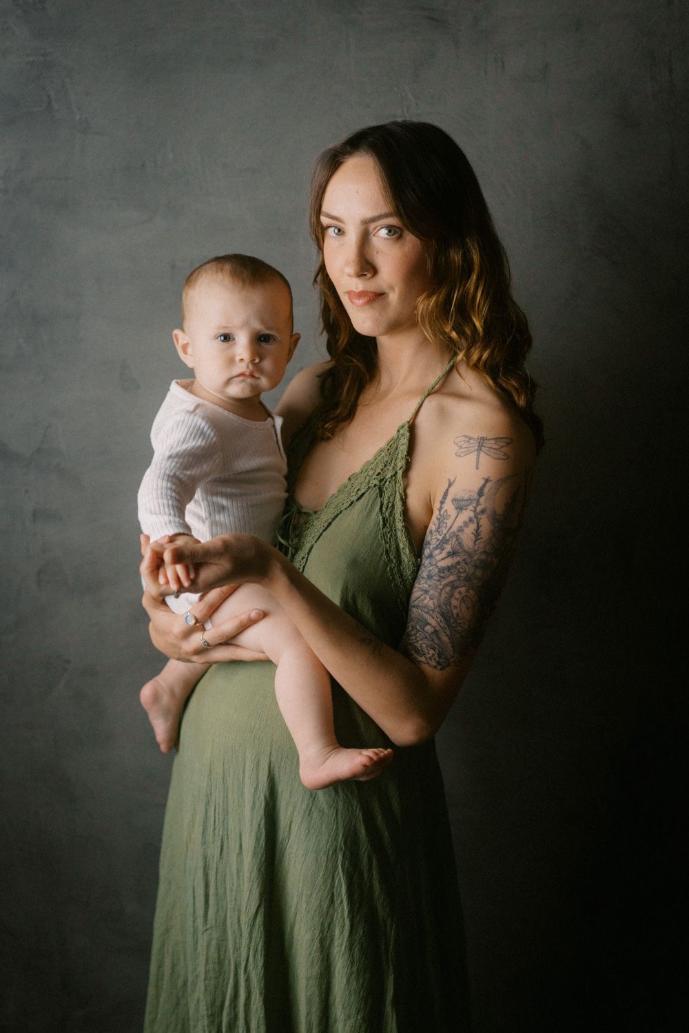 A tattooed mother in a green dress holds her baby in a studio portrait setting.