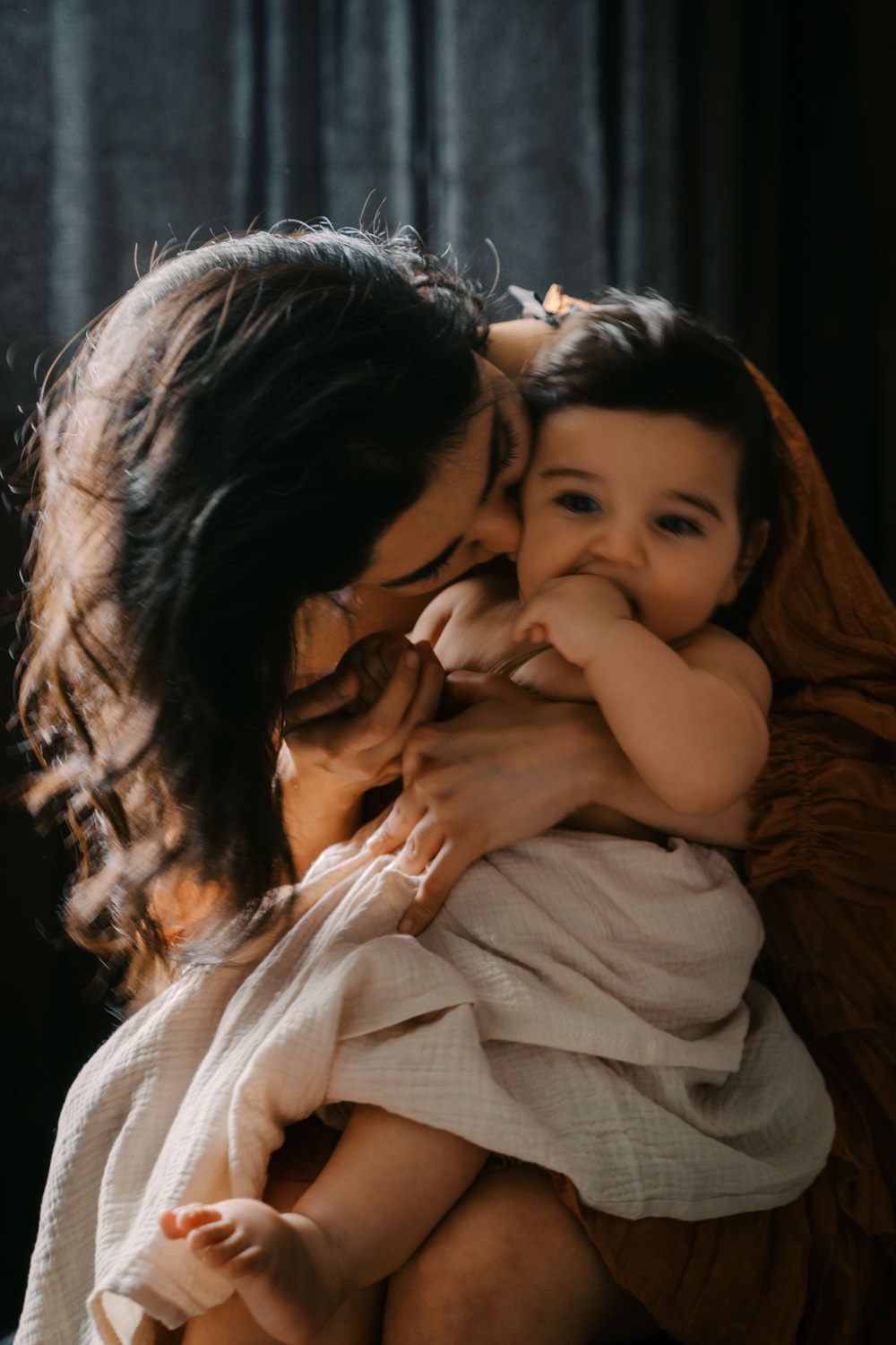 A mother tenderly kisses her smiling baby wrapped in a warm blanket in a softly lit portrait.