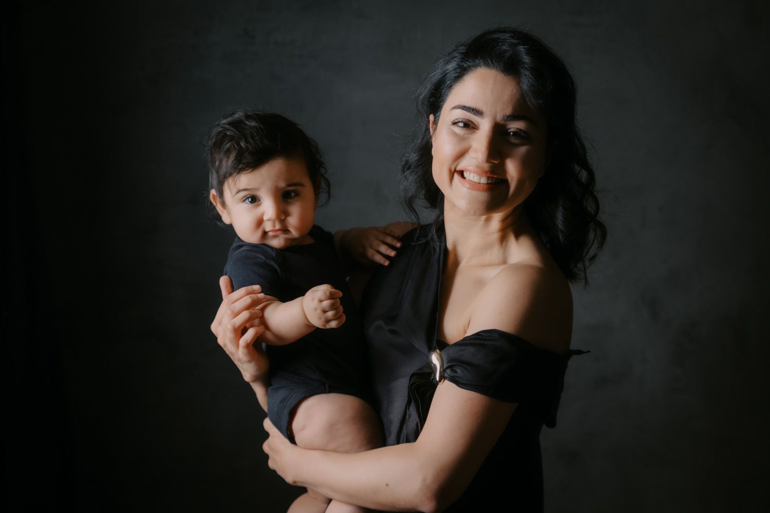 A smiling woman in a black off-shoulder top holds a baby dressed in black against a dark studio background.