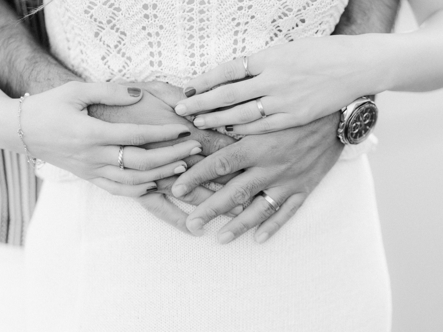 Black and white photo of couple holding hands over pregnant belly, wearing wedding rings and a watch.