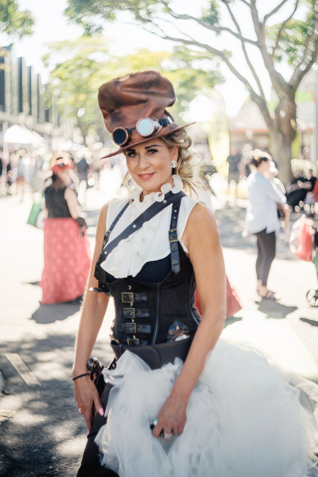Woman in steampunk costume with corset, top hat, and goggles at outdoor festival.