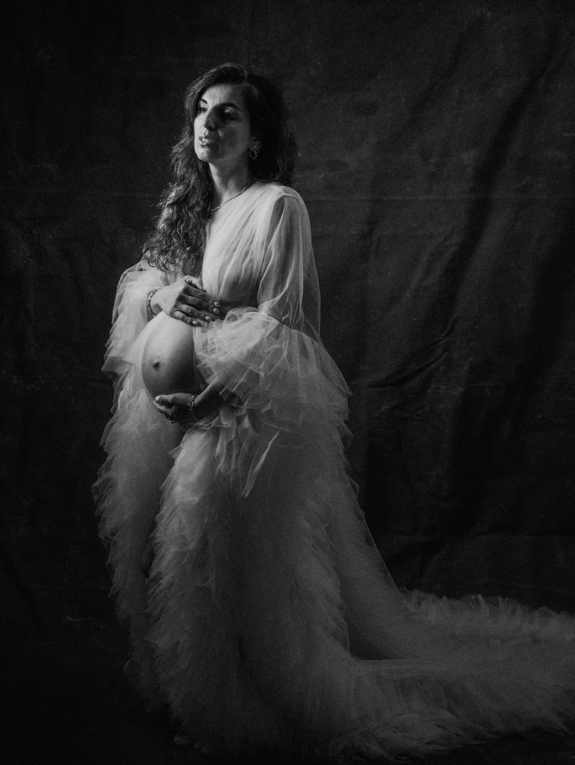 Black and white maternity portrait of a pregnant woman in a flowing white tulle gown, cradling her baby bump in a studio.