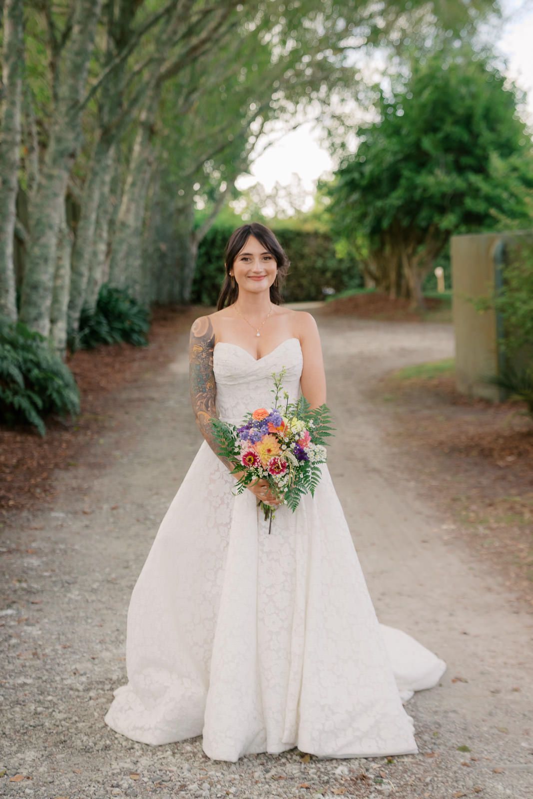 Bride in strapless lace wedding gown holding colorful bouquet stands on tree-lined path outdoors.