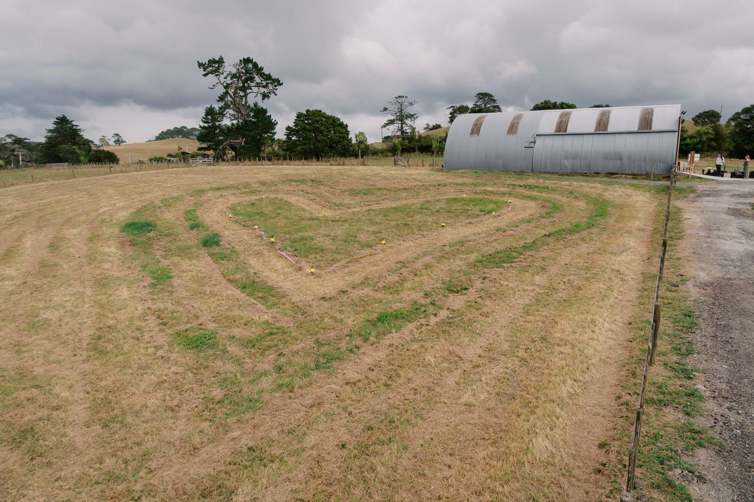 A large open field with mowed grass patterns and a corrugated metal barn under a cloudy sky.