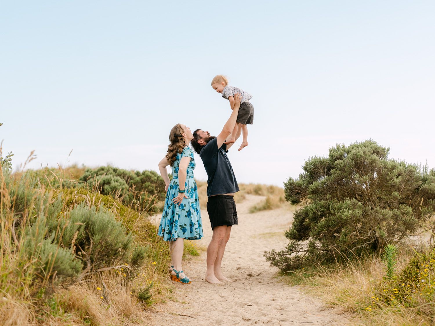 Family enjoying a sunny beach day, father lifting baby in the air while mother watches, surrounded by coastal dunes.