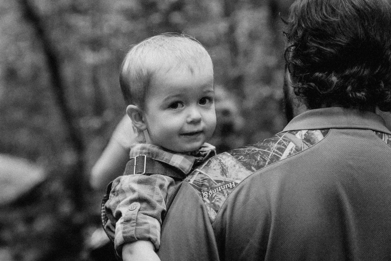 Black and white photo of a smiling toddler being carried on an adult's back outdoors in a wooded area.