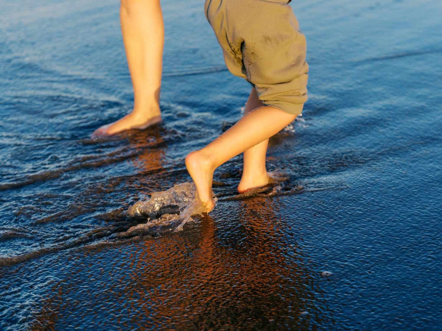Barefoot person walking in shallow ocean waves on a wet sandy beach at sunset wearing khaki shorts.