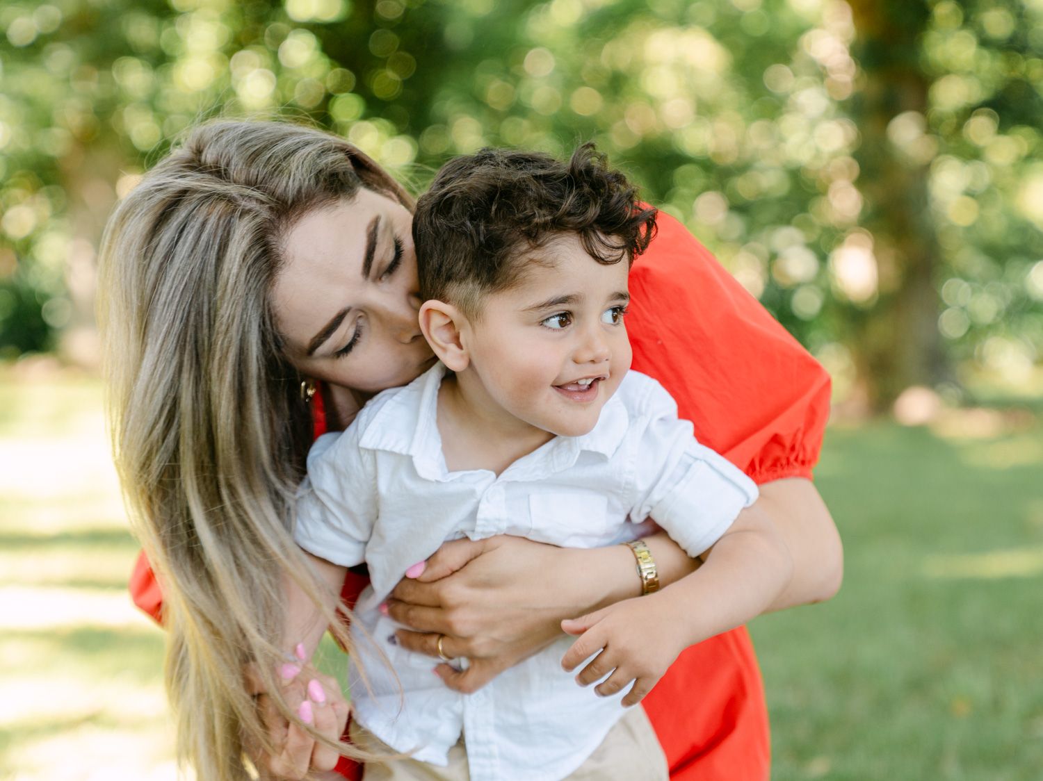 A loving mother in a red dress hugs and kisses her smiling toddler boy outdoors in a lush green park.