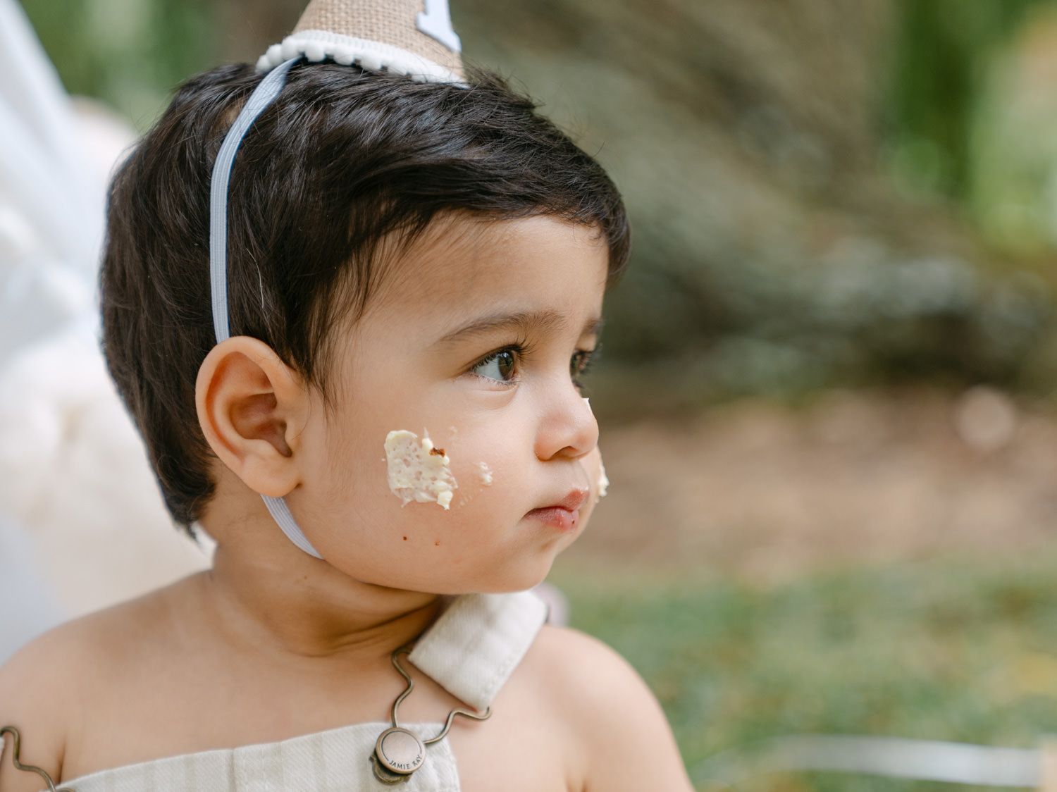 Cute toddler with cake on face wearing a white outfit and headband at an outdoor birthday party.