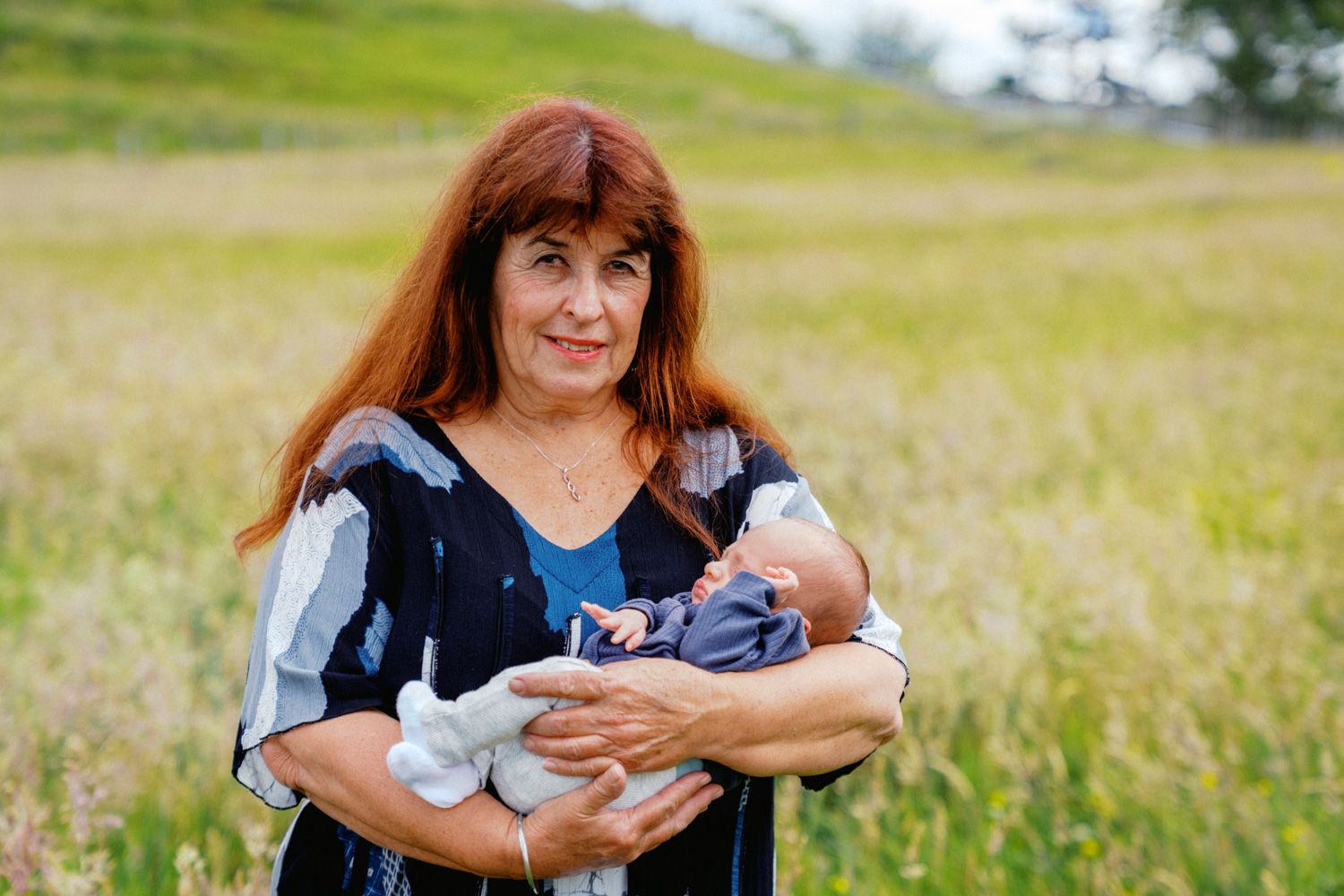 Woman with red hair holding a newborn baby in a field of green grass outdoors.