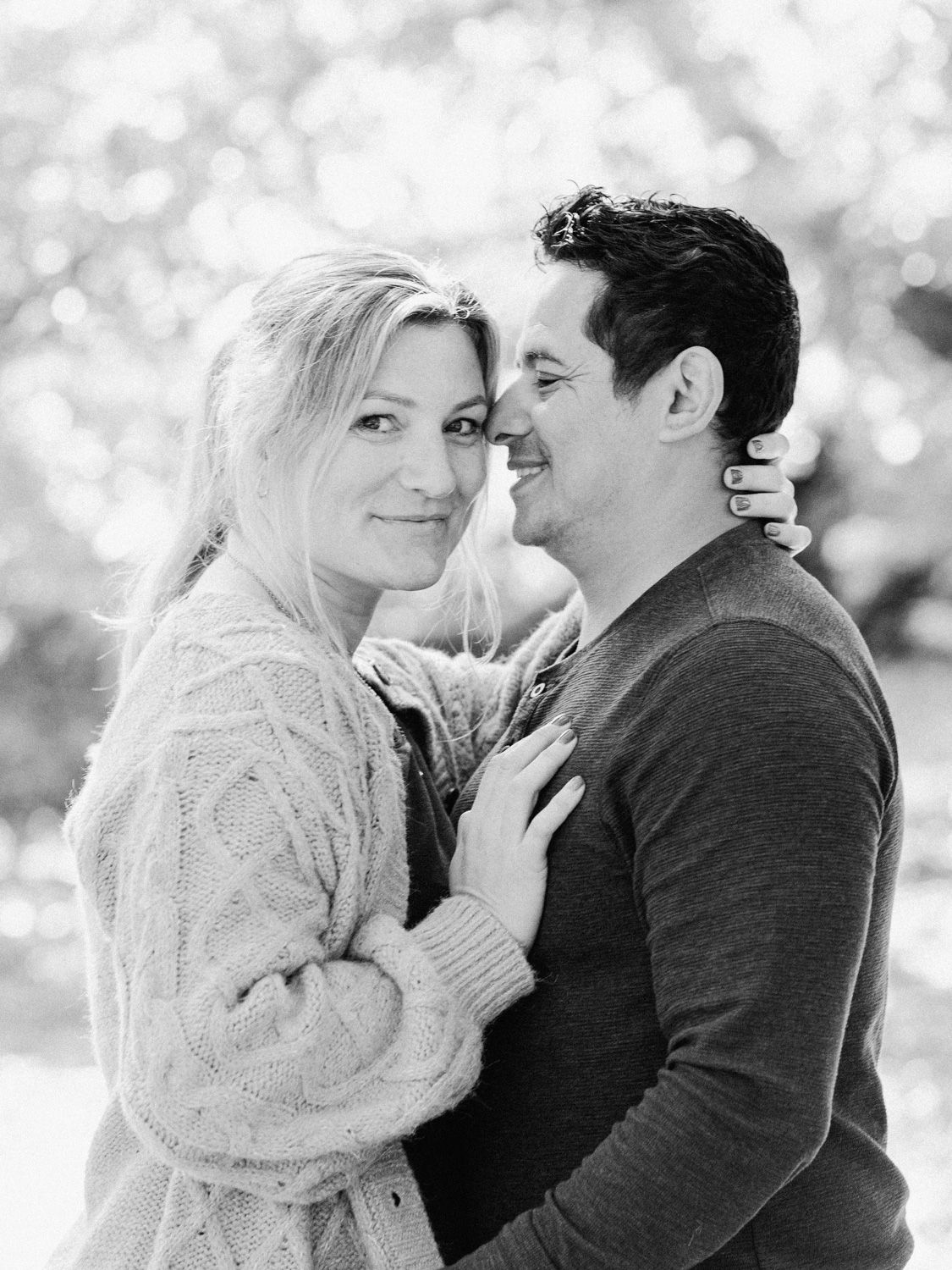 Black and white photo of a smiling couple embracing outdoors, woman in knit sweater resting head against man.