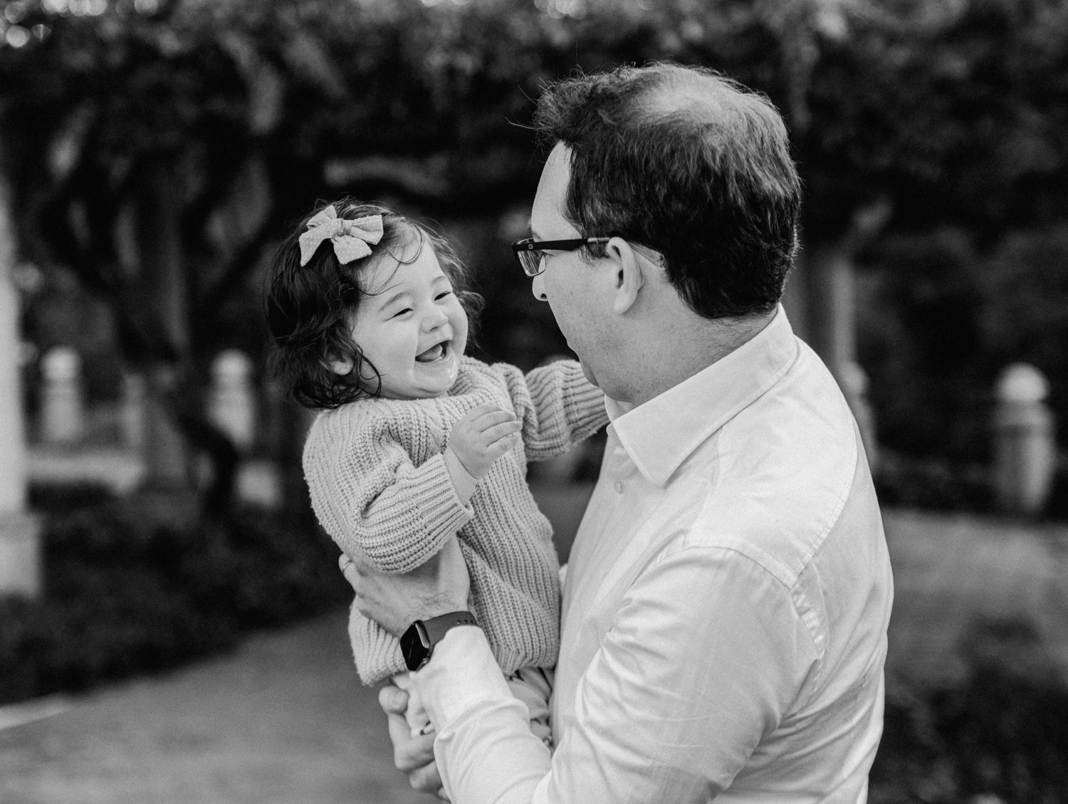Black and white photo of a man holding a laughing toddler girl with a bow in her hair outdoors.