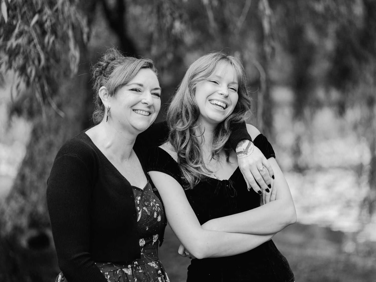 Two smiling women in black outfits pose together outdoors under willow trees in a black and white portrait photo.