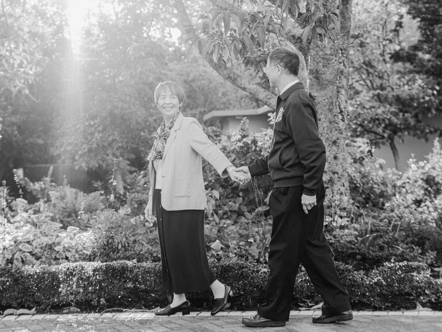 Black and white photo of a couple holding hands and walking through a lush garden setting.