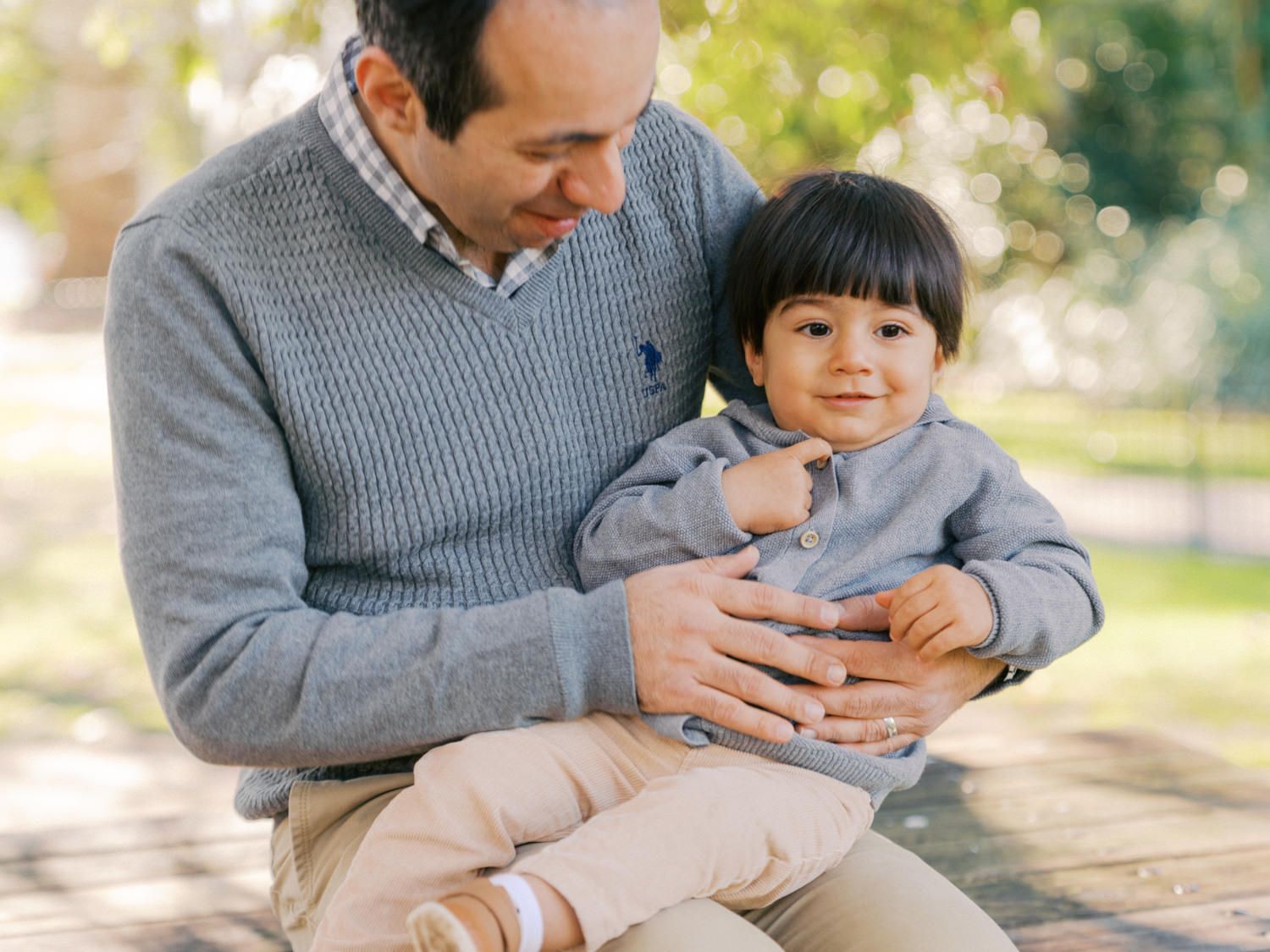 A father holds his young toddler son on his lap outdoors in a park, both wearing gray sweaters.